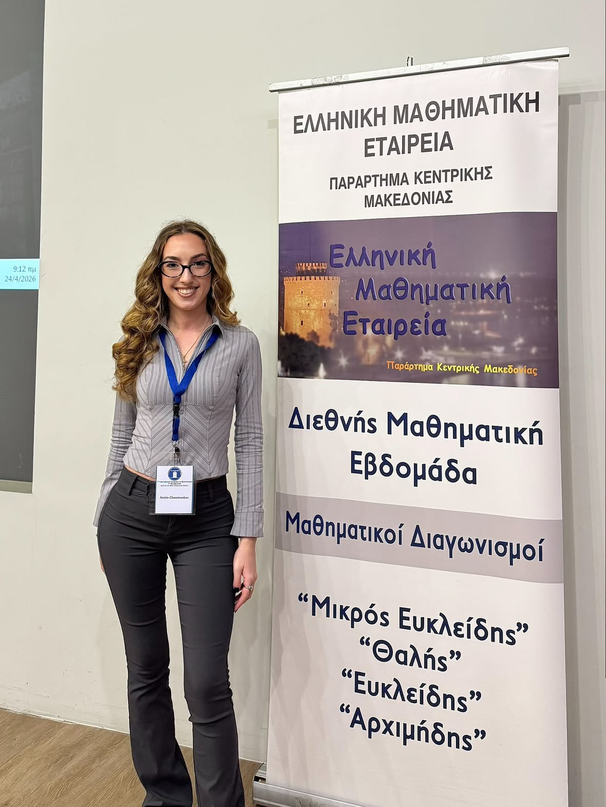 Sotiria Chatzimarkou at a Hellenic Mathematical Society event in Central Macedonia, standing beside a banner for International Mathematics Week